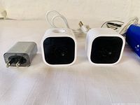 Two white Blink Mini security cameras side-by-side with attached white USB cables and a silver USB power adapter on a light surface.