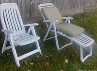 Two white foldable resin outdoor chairs on grass by a wooden fence. One is a straight armchair; the other is a lounge chair with beige cushion.