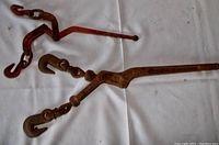 Two rusted metal load binders with grab hooks on both ends lying on a white cloth, showing the lever mechanism and hooks.