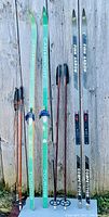 Photo showing two pairs of cross-country skis and two sets of ski poles leaning against a wooden background. Left pair is Can Sport touring skis in green and white with blue bindings. Right pair is Finn Arrow competition skate skis in gray with black and red bindings. Two sets of poles: thin black and orange, and black and red.