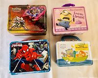 Four collectible tin lunch boxes from above showing front artwork including Pokémon, Minions, Spider-Man, and a cookie-themed design