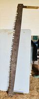 Full view of the antique crosscut saw showing metal blade with H-pattern teeth and oak handle at one end, placed on a white table.