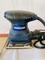 Close-up photo of the Mastercraft hand sander showing brand label, ergonomic design, and the sanding pad base.
