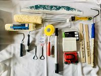 Full lot of painting supplies including roller covers, roller frames, stirring sticks, scraper, masking tape, and hand tools arranged on a white surface