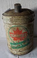 Image showing front side of vintage Supertest gas can with Canadian maple leaf logo and text 'SUPERTEST' and 'CANADA'S FINEST'. Rusted metal surface with wear.