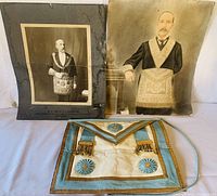 Two vintage black and white photographs of Masonic lodge members standing and wearing aprons, displayed behind a vintage Masonic apron with blue trim and gold details lying flat.