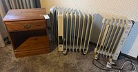 Photo of vintage wooden bedside table next to two oil-filled radiators and one ceramic panel heater on carpeted floor.