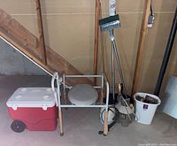 Full lot photo showing the raised toilet seat frame, cooler, broom, mop, walking cane, white plastic cane, and bucket with cleaning tools arranged against a basement wall.