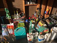 Wide view of gardening supplies including a new garden hose in packaging, bags of seed and fertilizer, spray bottles, watering cans, and various garden chemicals in plastic containers and metal cans.