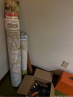 Three rolled vintage maps and a partially open box containing tools and accessories on the floor near a wall and electrical outlet.