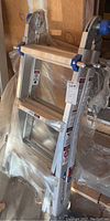 Folded aluminum Werner ladder with blue plastic parts in original plastic packaging, leaning against a wood wall.
