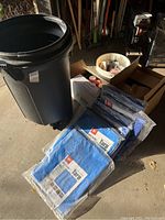 Grey plastic trash bin, six blue packaged tarps, boxes containing spray paint cans and miscellaneous items in garage setting.