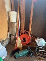 Corner setup with brooms, Orbit sprinkler, orange extension cord reel, bucket, and other gardening supplies