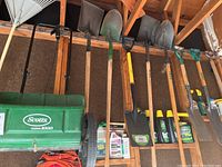 Photo of shovels, seed spreader, and rakes hanging on garage wall with some gardening supplies below