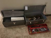 Two metal tool boxes, one with multiple hand tools organized in a red tote tray inside, one smaller unopened tool box with rolled chart inside.