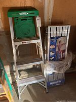 Front view of Werner aluminum stepladder with green top and plastic tool tray alongside packaged metal work platform, both stored in a garage.