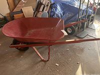 Red metal wheelbarrow side view showing tray, one wheel, and handles inside garage.