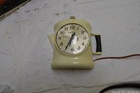 Front view of beige coffee pot shaped wall clock with round clock face and black handle, showing Arabic numeral dial and clock hands.