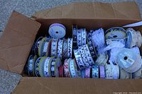 Open cardboard box containing numerous ribbon spools with various patterns and colors including animal and skeleton themes