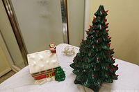 Photo showing ceramic Christmas tree and gingerbread house on a white cloth-covered surface.