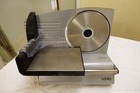 Front view of the stainless steel Cooks meat slicer showing circular blade, food holder with grip handle, and smooth slicing platform surface.