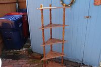 Full view of the oak corner shelf with four triangular shelves placed outdoors against a blue wall showing the entire structure including turned spindle supports and finials.