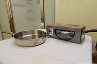 Frying pans displayed on table; one stainless steel pan out of box, one boxed with black handle visible.