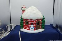 Front view of lighted ceramic house showing red brick pattern, snow-covered roof, Santa on chimney, snowman in front, and electrical cord.