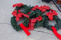 Five green artificial pine wreaths on ground, each decorated with red bows and long red ribbons, arranged side by side.