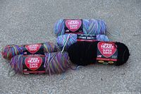 Five skeins of Red Heart Super Saver yarn lying on the ground: three variegated multi-color skeins with purple, blue, pink, yellow, and green tones, and two solid black skeins.