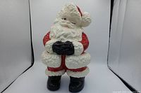Front view of ceramic Santa Claus figurine with red suit, white fur trim, black gloves and boots, winking eye, folded arms.