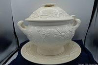 Off white soup tureen with floral embossed design, two handles, and matching underplate.