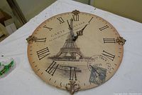 Top angled view of the round Paris themed clock showing the Roman numerals, Eiffel Tower illustration, and label details on a light colored surface.