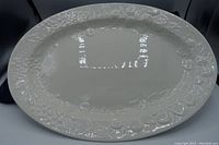 White oval ceramic serving platter with embossed floral border design on a black background, showing full top view.