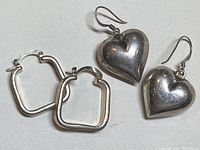 Two pairs of silver earrings; one pair heart-shaped drop earrings, one pair small square hoop earrings.