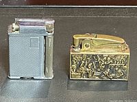 Two vintage lighters placed side by side on a dark surface; one silver textured rectangular lighter and one gold tone lighter with detailed relief artwork