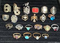 Photo shows 22 assorted costume rings arranged in four rows on a black display tray. Varied designs include floral, geometric, and gemstone styles in silver and gold-tone metals.