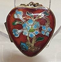 Front view of heart-shaped brass pill box with floral enamel decoration on reddish background.