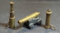 Three items on dark surface: two brass telescopes keychains and one brass naval cannon model with cast iron carriage.