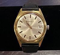 Front view showing gold-tone watch case, silver dial, black hour markers, date window, and black leather strap.