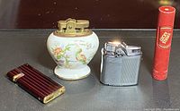 Front view of four assorted vintage lighters including Ronson Cobra, ceramic floral table lighter, metal lighter, and Lucky Strike container.