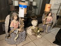 Photo shows two ceramic angel candle holders holding large colored candles and a ceramic sitting angel figurine with flowers, all on tiled floor.
