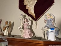 Three angel figurines displayed on a shelf under a wall hanging, featuring varying poses and colors.