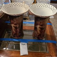Photo of two ceramic vases with cherub images and white lattice rims paired side by side on a glass table.