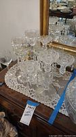 Two glass candelabras with hanging crystal pendants, two glass candle holders, two etched wine glasses labeled 'Nona' and 'Bill', arranged on a lace doily over a wooden table in front of a mirror.