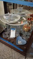 Wide angle photo showing porcelain service plate with floral pattern, teacups, crystal candle holders, metal trinket box, and other decorative dishes inside glass cabinet.