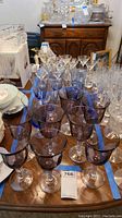 Photo showing multiple purple tinted goblet glasses and some clear glasses placed on a table, with wooden furniture and other glassware in the background.