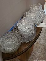 Seven matching crystal bowls stacked and arranged with two crystal serving platters underneath on a wooden table, showing intricate etched patterns and heavy cuts.