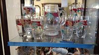 Glass pitcher and eight glasses on a glass shelf. The glasses are decorated with red flowers and green leaves.