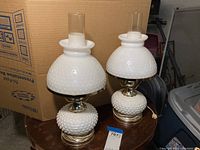 Pair of white hobnail glass lamps with textured broad bases and matching hobnail glass hurricane shades shown on a round wooden table.